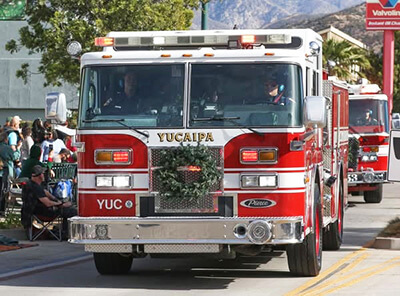 A Yucaipa Fire Department fire engine decorated with a holiday wreath drives in the Yucaipa Christmas Parade as spectators watch from the sidewalk.